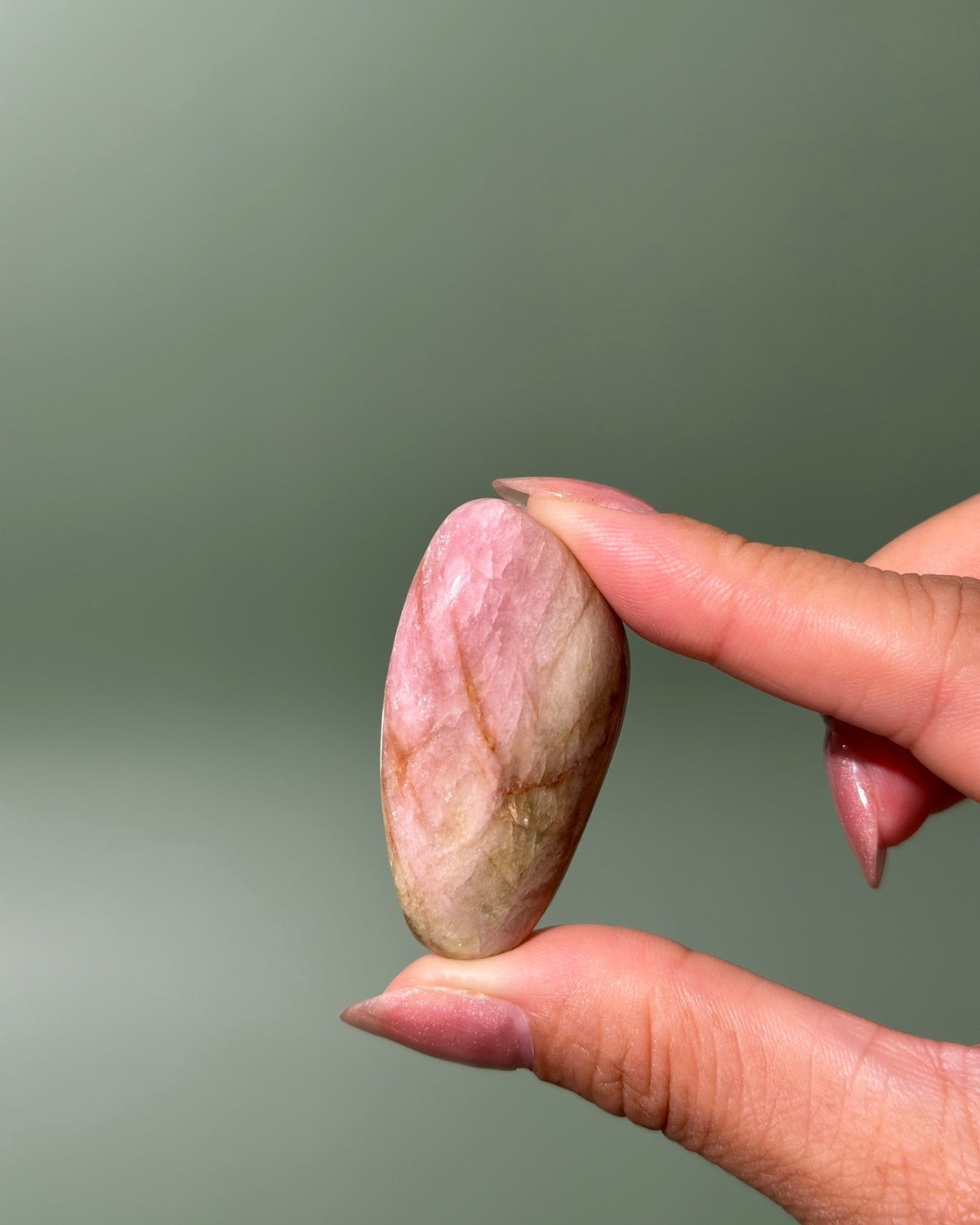 A hand with manicured nails holds a polished, oval-shaped pink stone with natural beige and brown veining against a soft, solid sage green background.