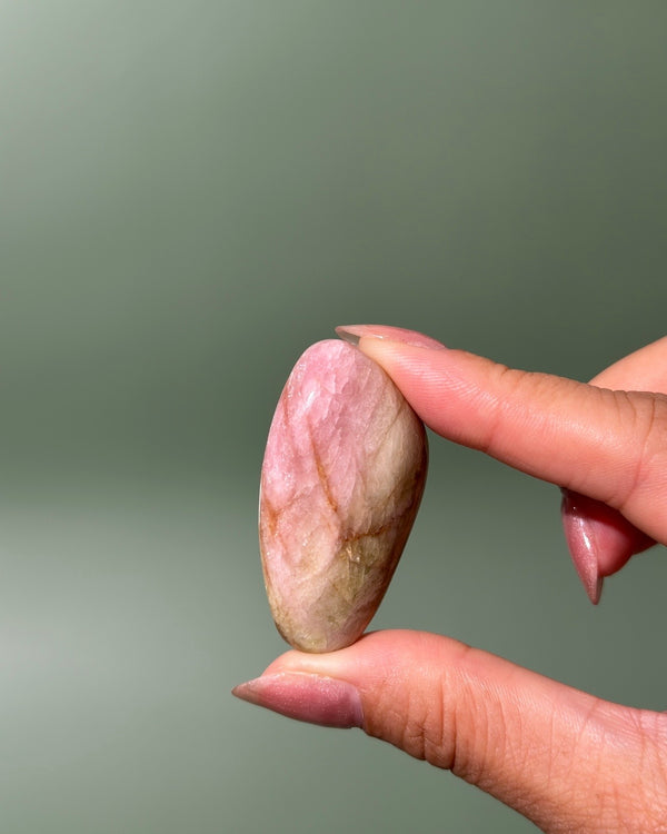 A hand with manicured nails holds a polished, oval-shaped pink stone with natural beige and brown veining against a soft, solid sage green background.
