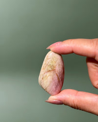 A hand holding a smooth, polished peach-colored stone with pink and cream marbling against a solid muted green background.