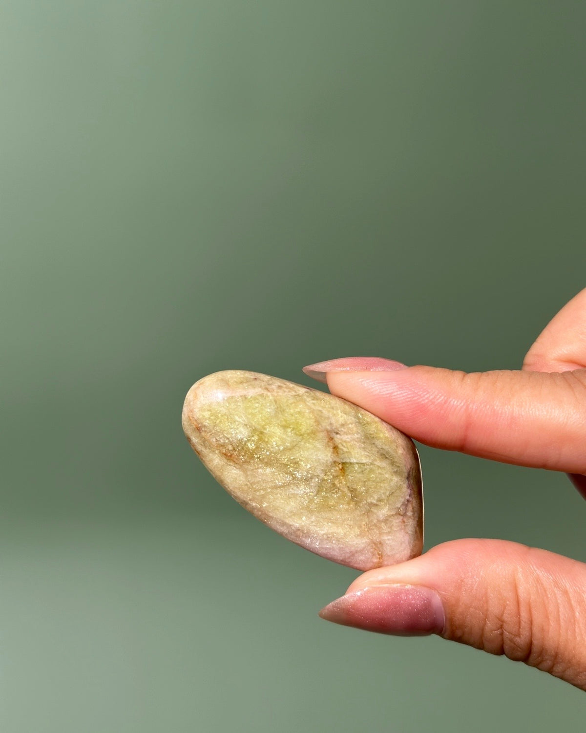 A hand holding a smooth, polished oval stone with natural beige, green, and pink yellow marbled patterns against a soft, solid sage green background.
