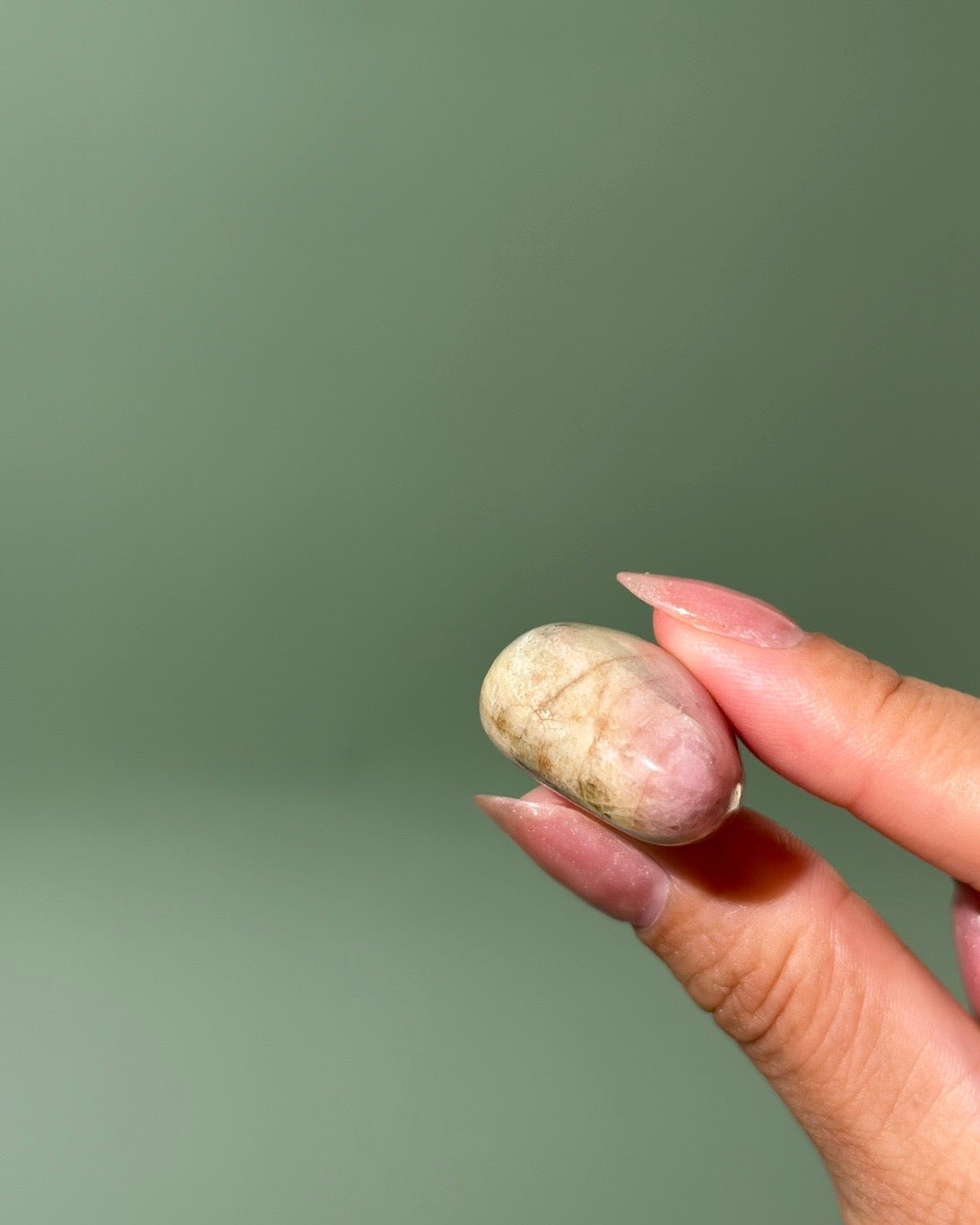A close-up of a hand holding a small, smooth, polished stone or bead with natural beige and light brown marbling against a solid muted sage green background.
