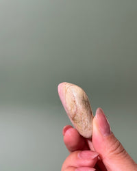 A hand with manicured nails holds a small, smooth, teardrop-shaped stone or crystal with a pinkish-beige and cream marbled texture against a solid, muted gray-green background.